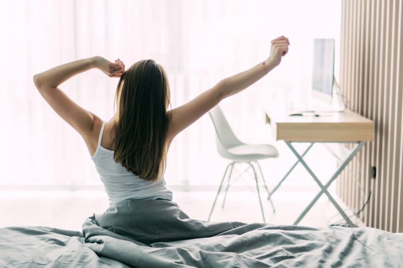 Mujer estirándose en la cama después de una noche de descanso reparador en un colchón HR de alta resiliencia, en una habitación moderna y luminosa.